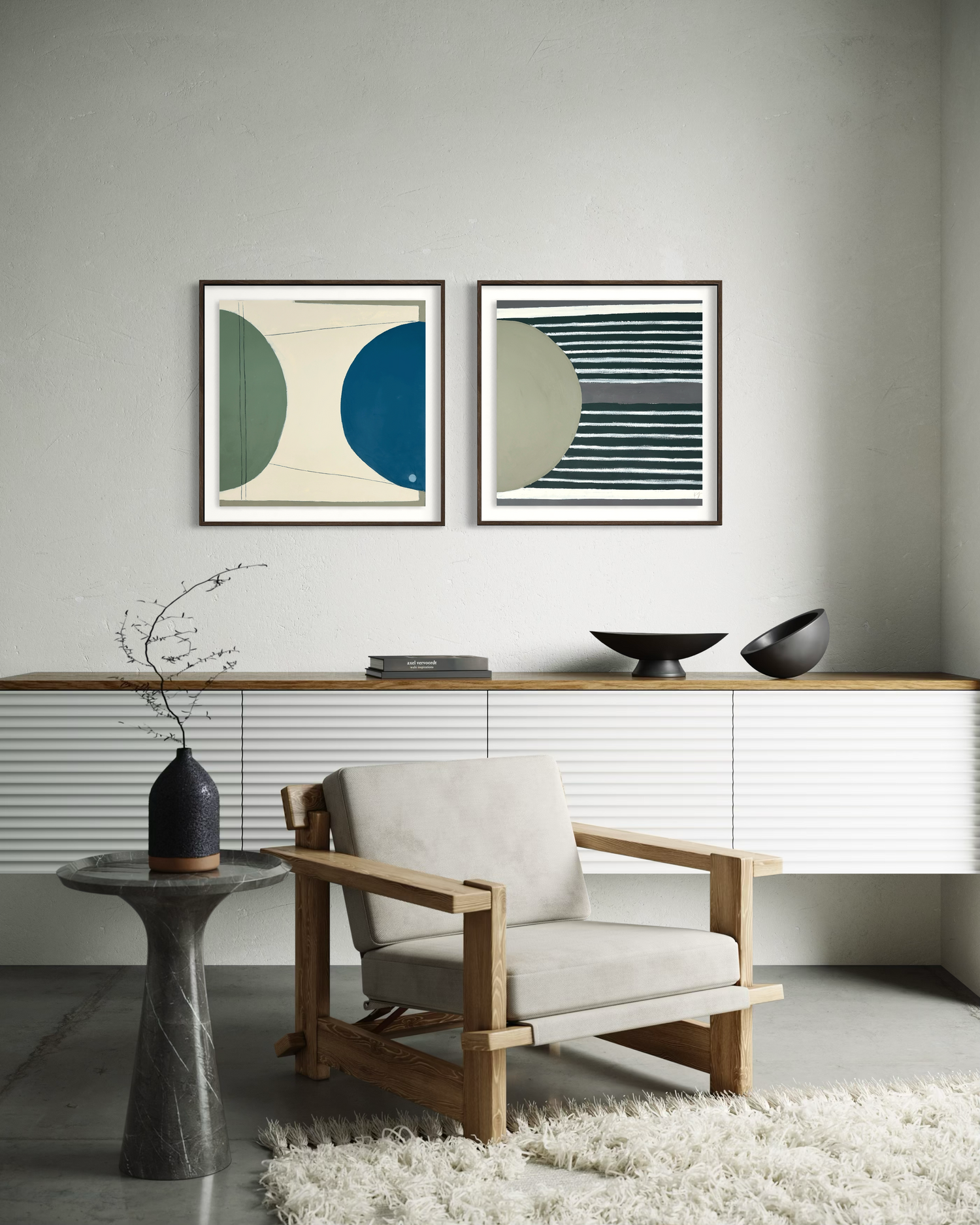 Two framed abstract prints hang above a wood bench, with a black bowl-shaped sculpture, vase, and cream chair against a light wall.